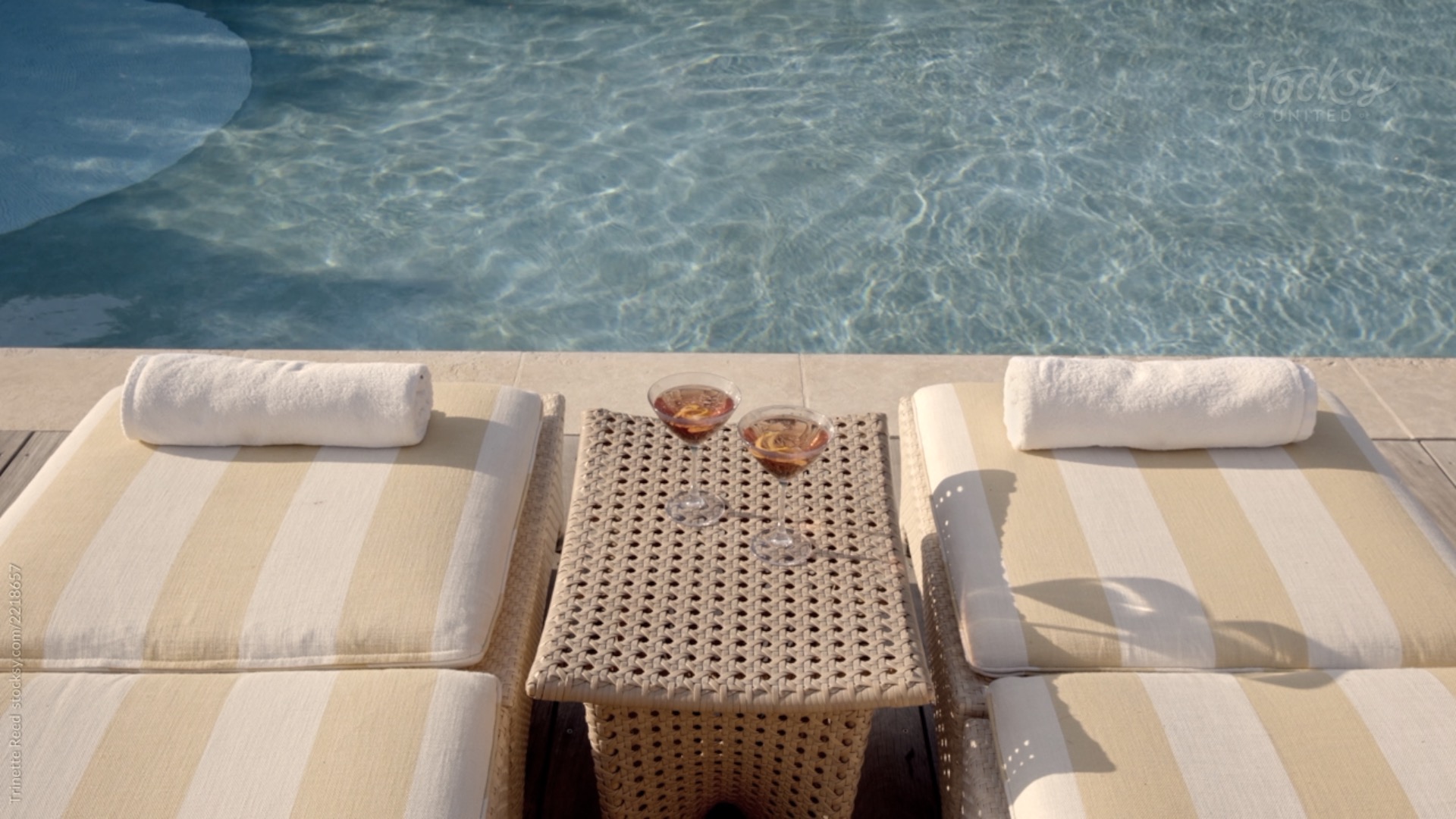 Aufnahme von zwei Liegen, geistreiften Liegenauflagen, zwei eingerollten Handtüchern und zwei Cocktails auf Tisch, im Hintergrund: Thermalwasser