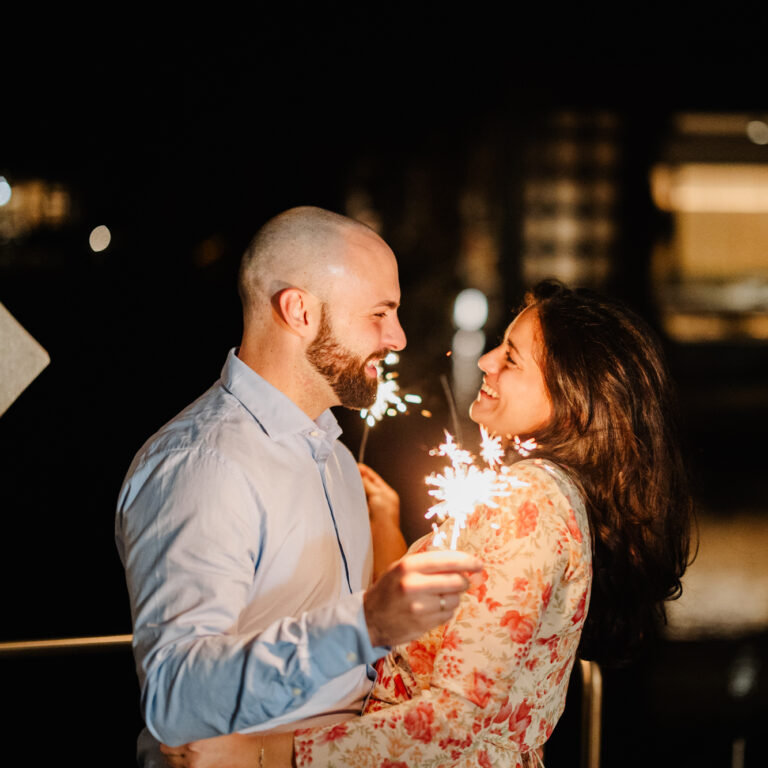 Pärchen steht auf der Terrasse in den Silent Villas in der Nacht, blickt sich an und lächelt und hält Sprühkerze in der Hand
