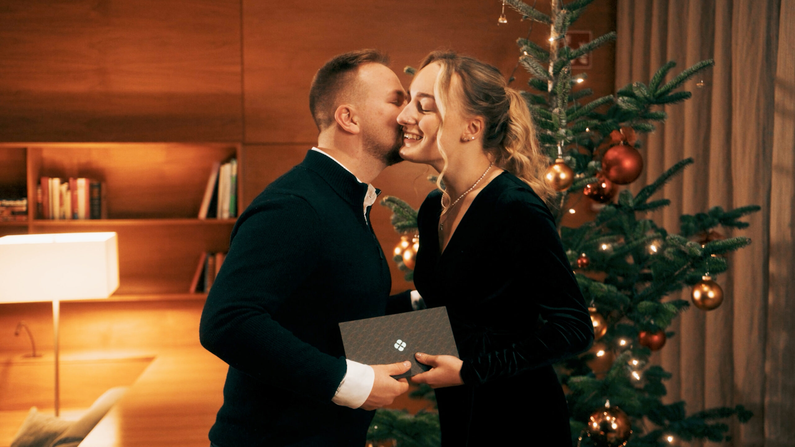 Junges Pärchen steht vor dem Christbaum und überreicht sich Gutschein